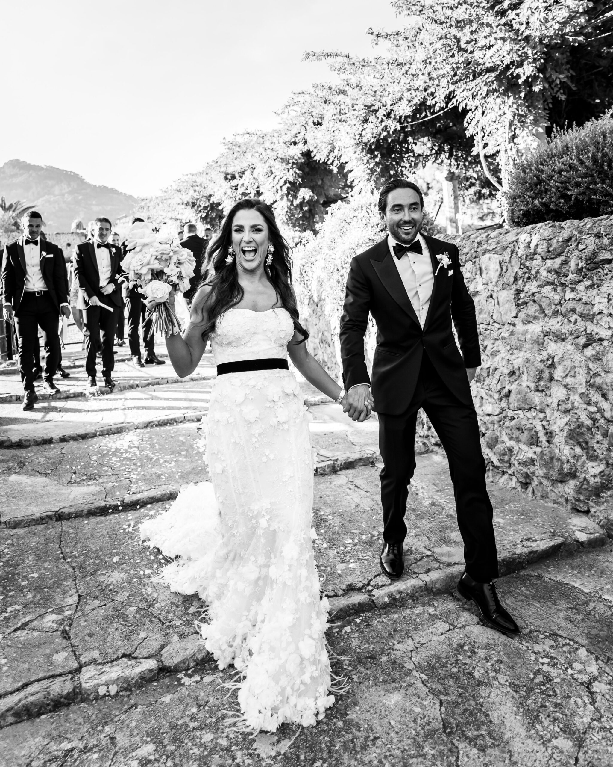 A couple walking through the gardens of Jardines De Alfabia in Mallorca after just being married, black and white stylish couple walking together on their wedding day, holding flower in the air celebrating their nuptials